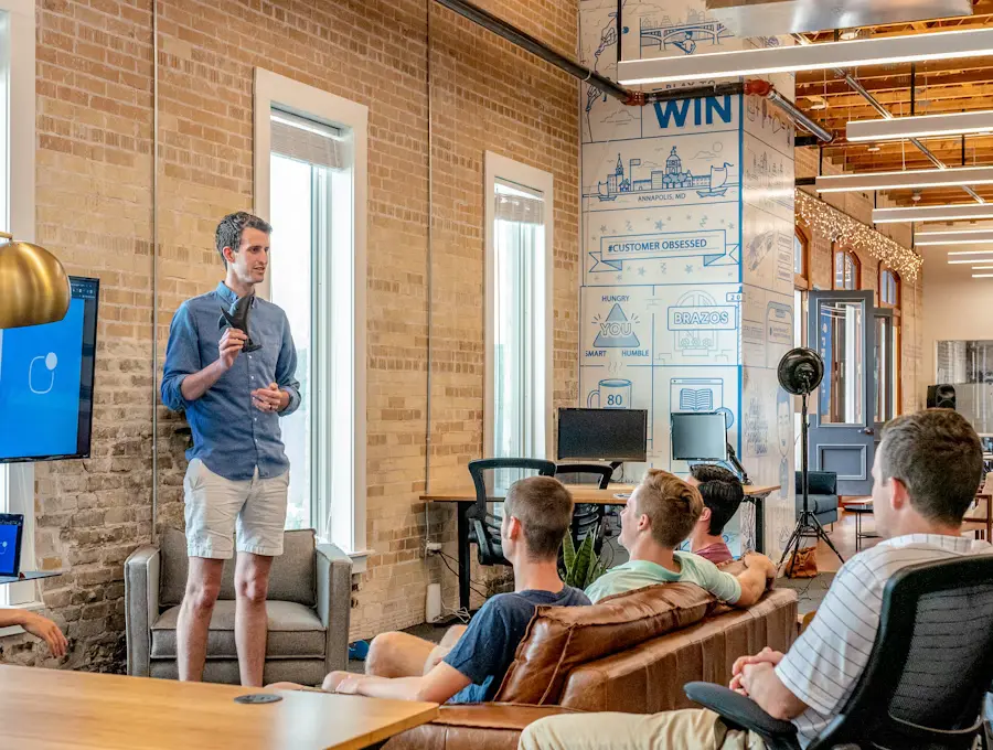 Man in blue shirt and white shorts presents to a small audience in a brick-walled coworking space, gesturing with a microphone.